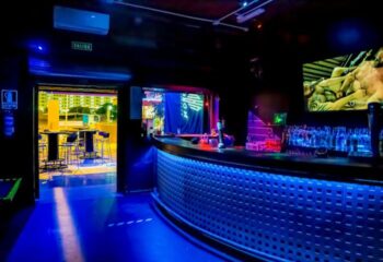 Interior of Noxon Club, a gay cruise club in Gran Canaria. Blue lighting illuminates the bar and dance floor. Tables visible outside.