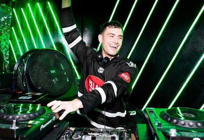 DJ at CIRCA Soho, gay bar in London's Old Compton Street. Man smiles, wearing hockey jersey, at decks with green neon lights.