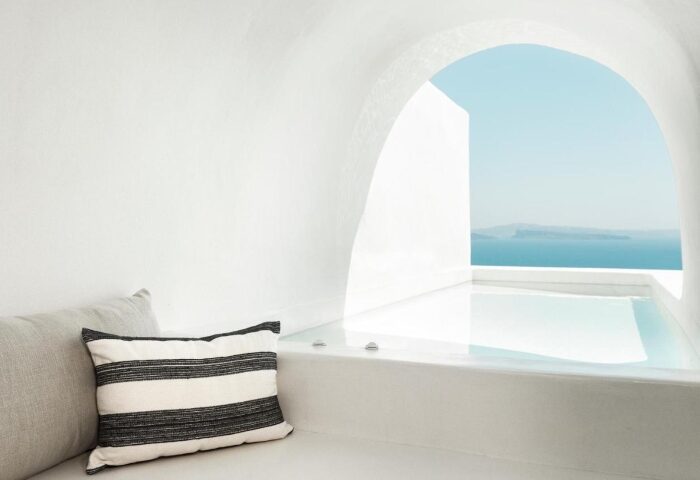 Armenaki Santorini hotel rooftop pool view. Gay hotels list travel guide. White architecture, blue sea, striped pillow. Gay travel.
