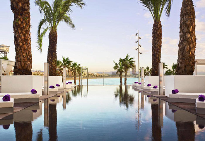 W Barcelona beachfront gay hotel pool view. Palm trees, sunbeds, and ocean backdrop. Number one gay hotel in Barcelona for gay men travel.