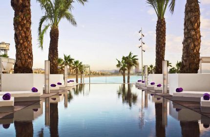 W Barcelona beachfront gay hotel pool view. Palm trees, sunbeds, and ocean backdrop. Number one gay hotel in Barcelona for gay men travel.