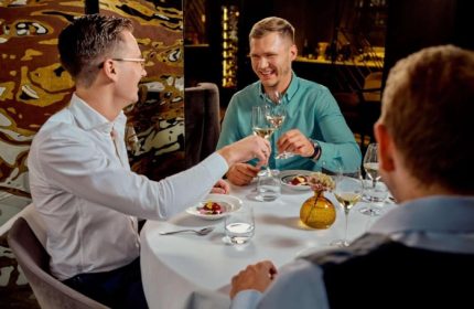 Gay honeymoon at Sofitel Legend The Grand Amsterdam. Three men toasting with white wine at a table in a restaurant. Gay travel in Amsterdam.