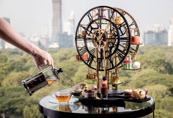 Afternoon tea in Bangkok's gayborhood, Silom. A hand pours tea near a Ferris wheel display of pastries. Rooftop view. Gay travel.