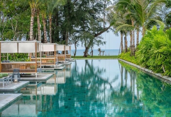 Melia Phuket Mai Khao, a wellness gay beach resort. Cabanas line an infinity pool reflecting palm trees and the ocean. Serene, tropical luxury.