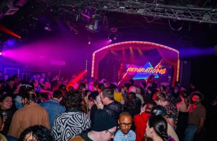 Oasis drag cabaret in San Francisco. A crowd of people at a drag show in a nightclub with a stage and pink and purple lighting. Gay travel.