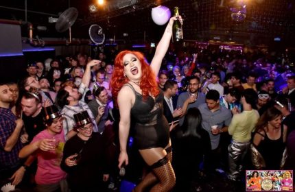 Oasis Drag Cabaret in San Francisco: A drag queen in lingerie and fishnets holds a bottle aloft in a crowded nightclub. Gay travel.