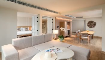 SAii Laguna Phuket ocean-front suite: living area with couch, table, and hat. Bedroom and dining area in background. Luxury gay travel.