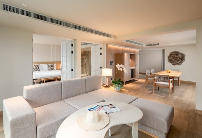 SAii Laguna Phuket ocean-front suite: living area with couch, table, and hat. Bedroom and dining area in background. Luxury gay travel.