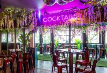 On Bar Manchester: Stylish interior with tables, chairs, plants, and a COCKTAILS neon sign. Gay travel advice.