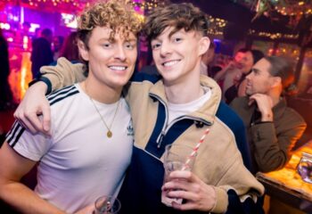 Two smiling gay men at On Bar, Manchester. One wears an Adidas tee, the other a color-blocked jacket. Both hold drinks in a lively bar scene.