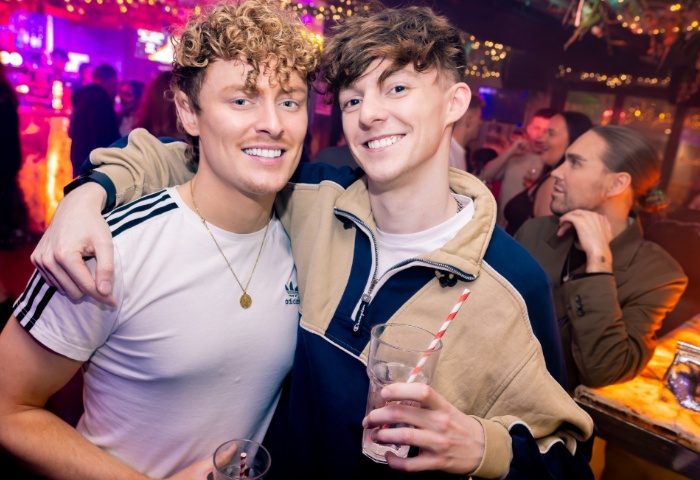 Two smiling gay men at On Bar, Manchester. One wears an Adidas tee, the other a color-blocked jacket. Both hold drinks in a lively bar scene.