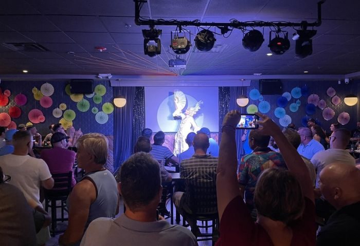 OneEleven Bar drag show. A drag queen performs on stage with a fan, illuminated by stage lights, as a crowd watches and takes photos.