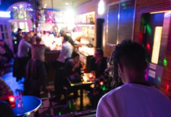 Oscar Bar Cafe Copenhagen scene. Gay bar interior with patrons at bar, tables, and DJ. Nightlife, travel advice.