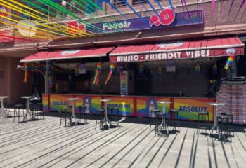 Parrots Pub, a gay bar in Yumbo Centre, Maspalomas, Gran Canaria. Rainbow decorations, bar tables, and Music-Friendly Vibes sign. Gay travel.