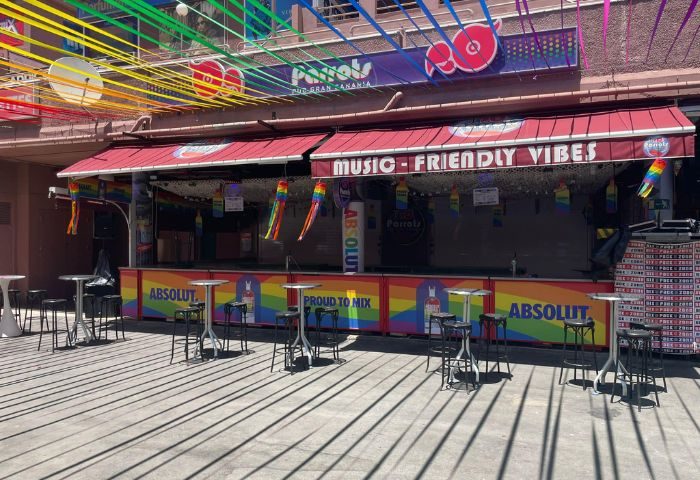 Parrots Pub, a gay bar in Yumbo Centre, Maspalomas, Gran Canaria. Rainbow decorations, bar tables, and Music-Friendly Vibes sign. Gay travel.
