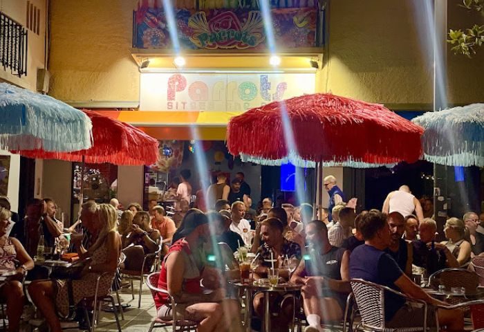 Parrots Pub terrace, a popular gay bar in Sitges, Spain. People sit at tables under colorful umbrellas. Nightlife scene.