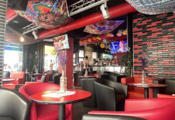 Parrots Pub terrace, a gay bar in Sitges. Interior view with red & black seating, TVs, and colorful decor. Gay travel destination.