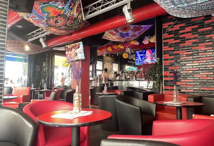 Parrots Pub terrace, a gay bar in Sitges. Interior view with red & black seating, TVs, and colorful decor. Gay travel destination.