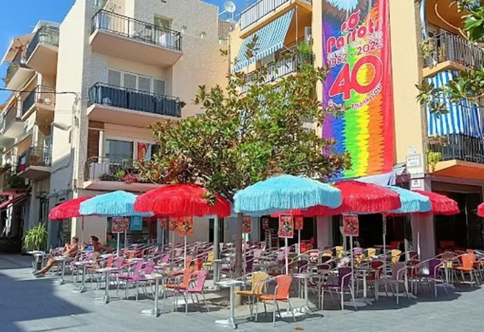 Parrots Pub terrace, a gay bar in Sitges. Colorful umbrellas shade tables and chairs. A rainbow banner celebrates the bar's 40th anniversary.