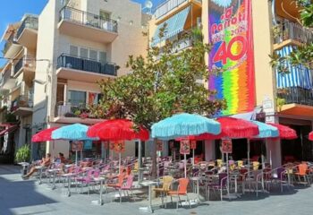 Parrots Pub terrace, a gay bar in Sitges. Colorful umbrellas shade tables and chairs. A rainbow banner celebrates the bar's 40th anniversary.