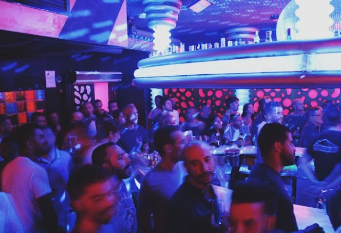 Inside Parthenon Gay Cruise Club in Torremolinos. Crowded bar scene with men under blue & pink lights. Gay nightlife travel.