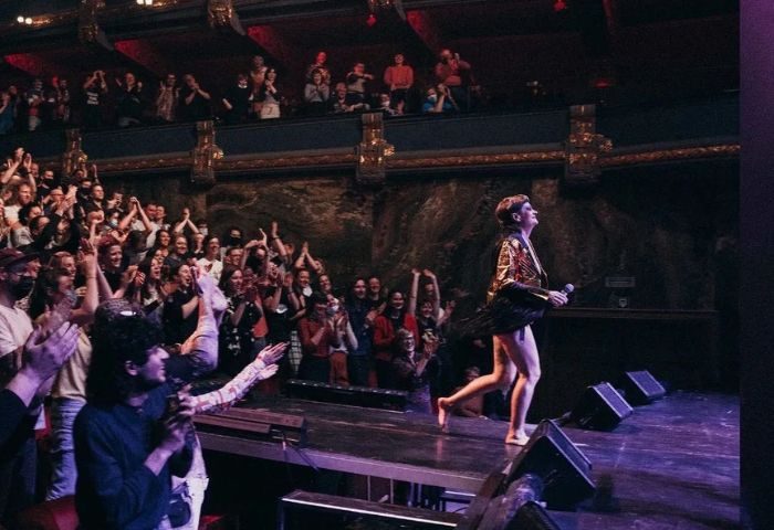 Peaches in Toronto concert. Singer on stage, barefoot, with a jacket, holding a microphone. Audience clapping. Gay travel advice.