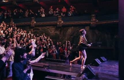 Peaches in Toronto concert. Singer on stage, barefoot, with a jacket, holding a microphone. Audience clapping. Gay travel advice.
