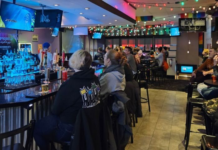 Interior view of Pegasus on Church, a gay bar in Toronto. People sit at the bar and tables, with TVs and colorful lights.