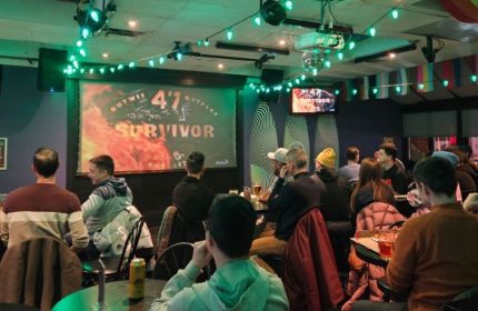Pegasus On Church, a gay bar in Toronto. Patrons watch Survivor on a large screen. Green lights decorate the ceiling. A fun gay travel spot.