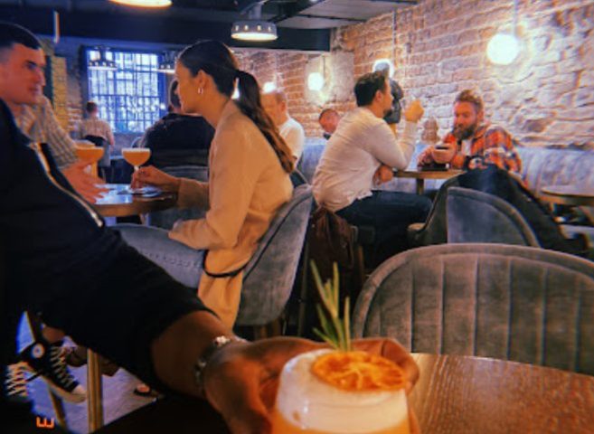 Patrons enjoy drinks at Penny Lane, a gay bar in Dublin. Brick walls, soft lighting, cocktails, and velvet seating create a cozy atmosphere.