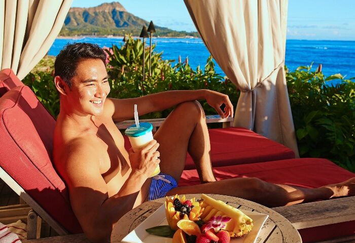 Gay group's perfect beach holiday at Sheraton Waikiki Beach Resort, Hawaii. Man relaxing with a drink and fruit, Diamond Head in background.
