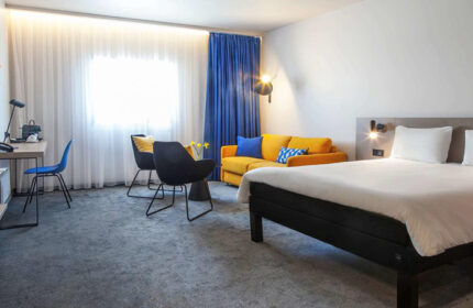 Ibis Praha Old Town Hotel room. Modern design with a bed, yellow sofa, desk, and chairs. Perfect for gay couples visiting Prague.