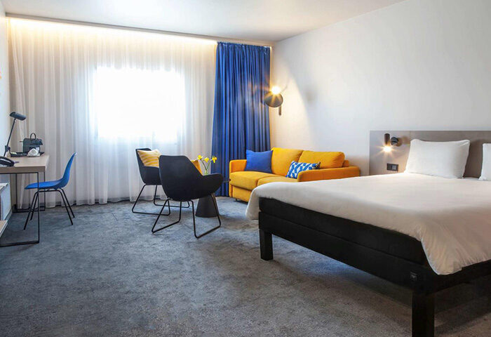 Ibis Praha Old Town Hotel room. Modern design with a bed, yellow sofa, desk, and chairs. Perfect for gay couples visiting Prague.