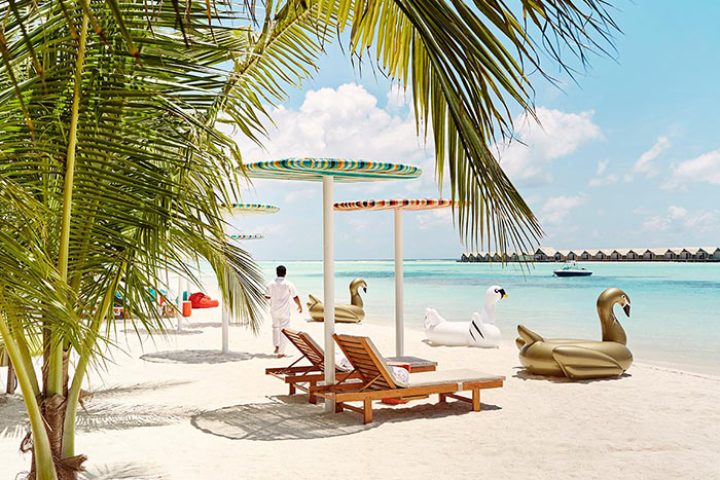 LUX* South Ari Atoll, Maldives beach. Lounge chairs, swan floats, and colorful umbrellas on white sand. Perfect gay honeymoon resort.