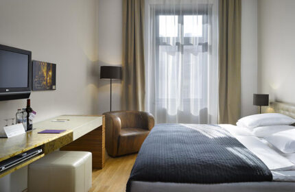 The Icon Hotel and Lounge room in Prague Old Town. Gay honeymoon hotel with bed, chair, desk, TV, wine, and window.