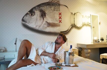 Gay traveler in Andaz Amsterdam Prinsengracht hotel room. Man in robe with champagne, fish/bottle art. Perfect Amsterdam hotel for gay solo travel.