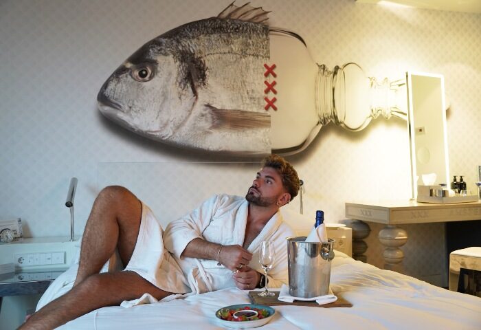 Gay traveler in Andaz Amsterdam Prinsengracht hotel room. Man in robe with champagne, fish/bottle art. Perfect Amsterdam hotel for gay solo travel.