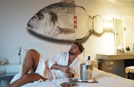 Gay traveler in Andaz Amsterdam Prinsengracht hotel room. Man in robe with champagne, fish/bottle art. Perfect Amsterdam hotel for gay solo travel.
