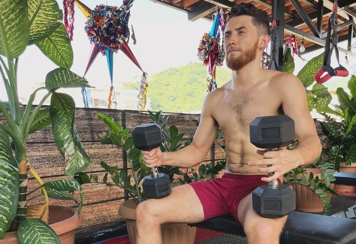Gay man lifting weights at Piñata PV Gay Hotel in Puerto Vallarta. He's shirtless, wearing red shorts, surrounded by plants and a piñata.