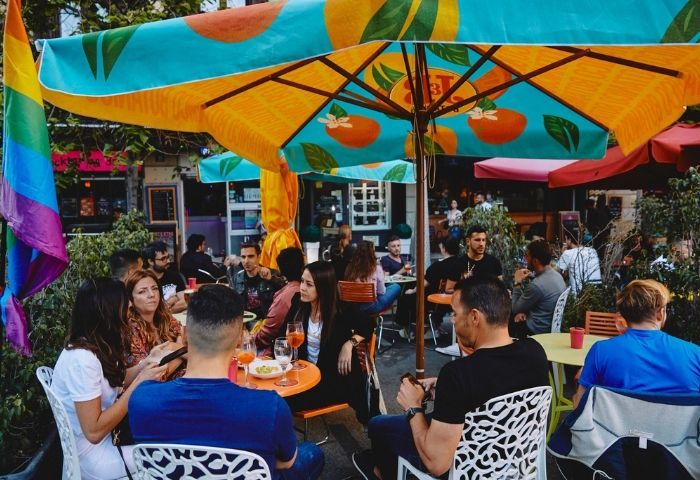 Gay bar in Barcelona: Outdoor seating at a pink corner bar. Rainbow flag visible, patrons enjoying drinks under colorful umbrellas.