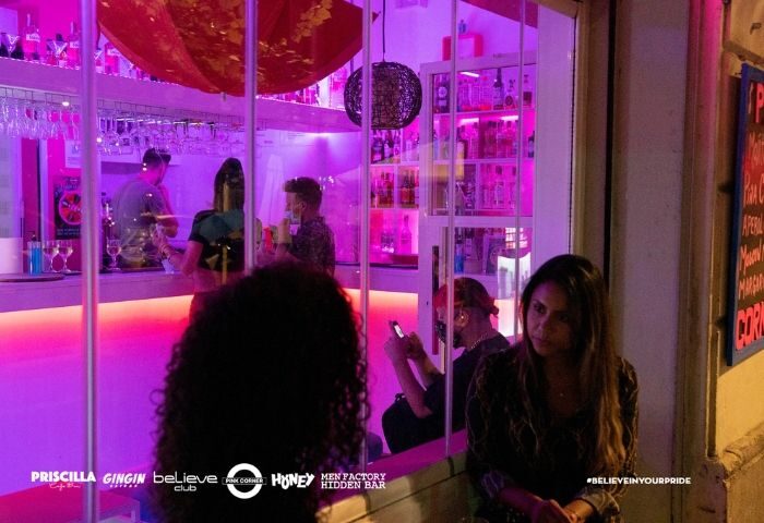 Gay Barcelona: Pink Corner gay bar's exterior, lit with pink and purple hues. People inside and outside the bar. #BELIEVEINYOURPRIDE.