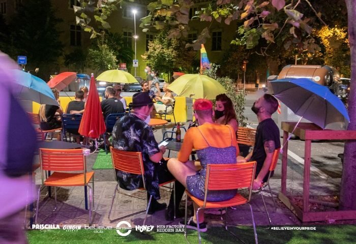 Outdoor seating at a pink corner gay bar in Barcelona. People sit at tables under umbrellas. The scene is lively and welcoming. Gay travel advice.