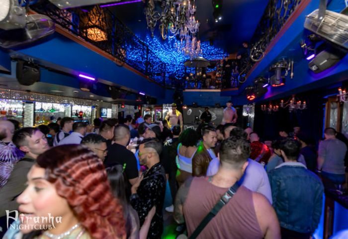 Inside Piranha Nightclub in Las Vegas, a popular gay bar. Patrons mingle under blue lights and chandeliers. Male dancer on stage.