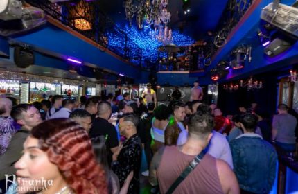 Inside Piranha Nightclub in Las Vegas, a popular gay bar. Patrons mingle under blue lights and chandeliers. Male dancer on stage.