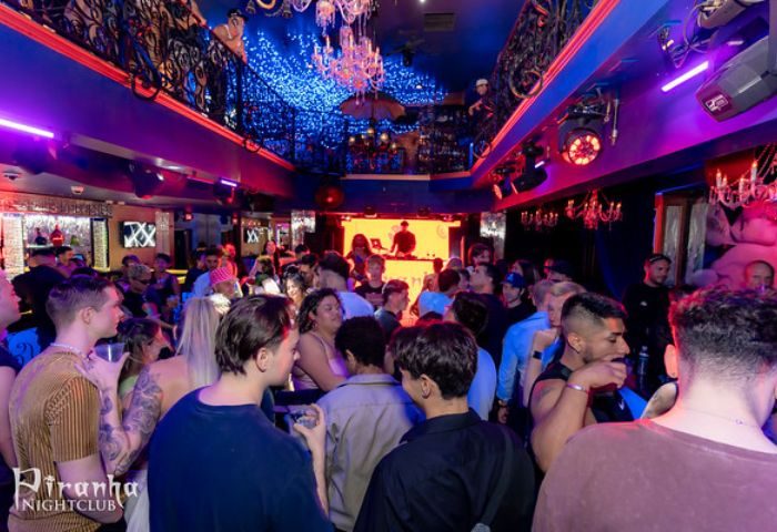 Inside Piranha Nightclub in Las Vegas. Crowd of gay men, DJ booth, chandeliers, and blue lighting create a vibrant atmosphere.