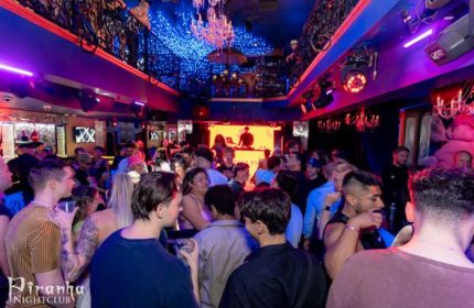 Inside Piranha Nightclub in Las Vegas. Crowd of gay men, DJ booth, chandeliers, and blue lighting create a vibrant atmosphere.