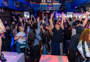 Inside Piranha Nightclub in Las Vegas, a crowd of gay men dance with their hands in the air under blue lights, enjoying the vibrant nightlife.