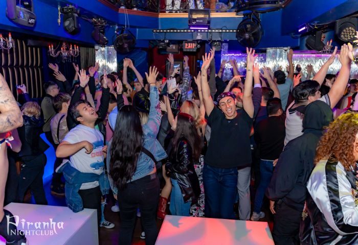 Inside Piranha Nightclub in Las Vegas, a crowd of gay men dance with their hands in the air under blue lights, enjoying the vibrant nightlife.