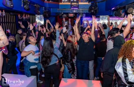 Inside Piranha Nightclub in Las Vegas, a crowd of gay men dance with their hands in the air under blue lights, enjoying the vibrant nightlife.