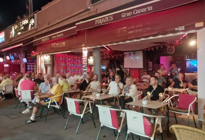 Patrons at Pirates Bar, a gay bar in Gran Canaria, enjoy drinks outside under the awning. Tables and chairs fill the outdoor seating area.